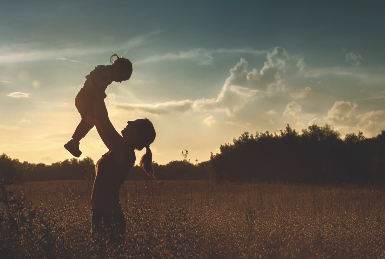 Madre alzando a su hija en brazos al atardecer en un campo, siluetas a contraluz