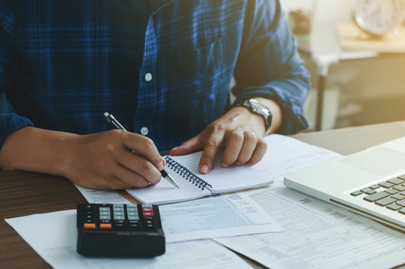 Persona revisando documentos y haciendo cálculos para la declaración de la renta junto a un portátil y una calculadora.