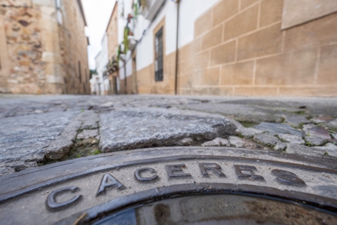 Detalle de una calle del casco antiguo de Cáceres con una tapa metálica en primer plano y edificios históricos al fondo.